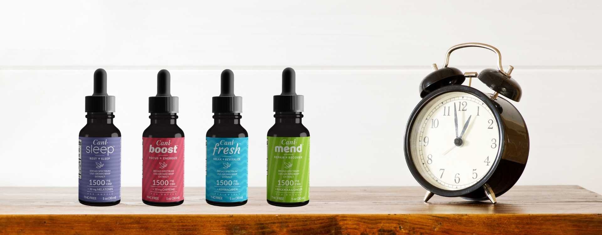 Alt=“Four CBD oil tinctures next to a black alarm clock placed on a wooden table”